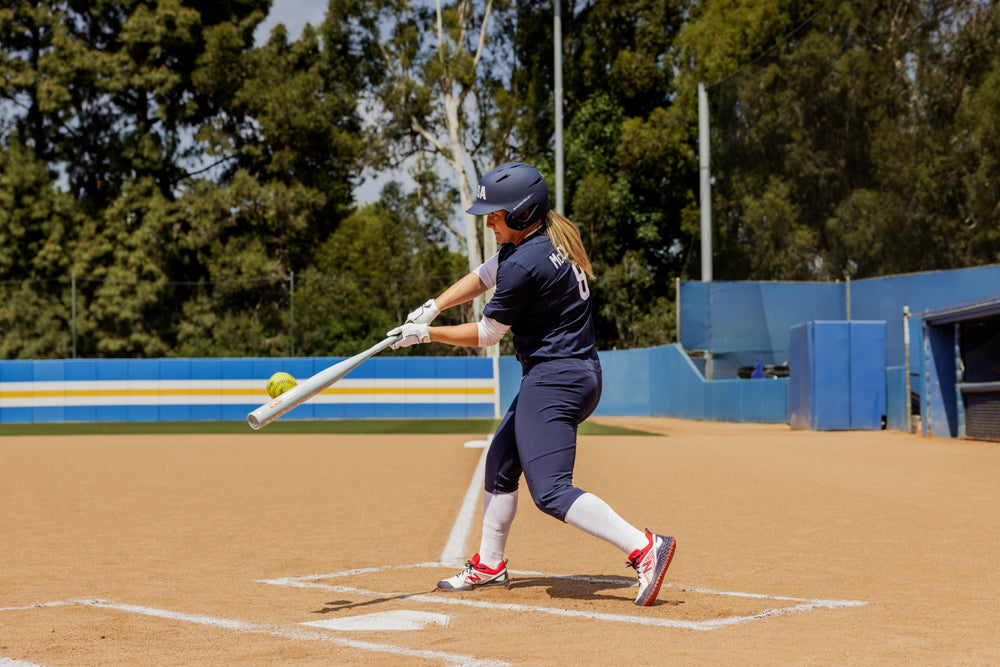 A softball player in navy swings the 2025 Easton Ghost Unlimited (-9) Fastpitch Softball Bat (EFP5GHUL9) by Easton, hitting a yellow softball at home plate on a dirt field with trees and a blue fence in the background.