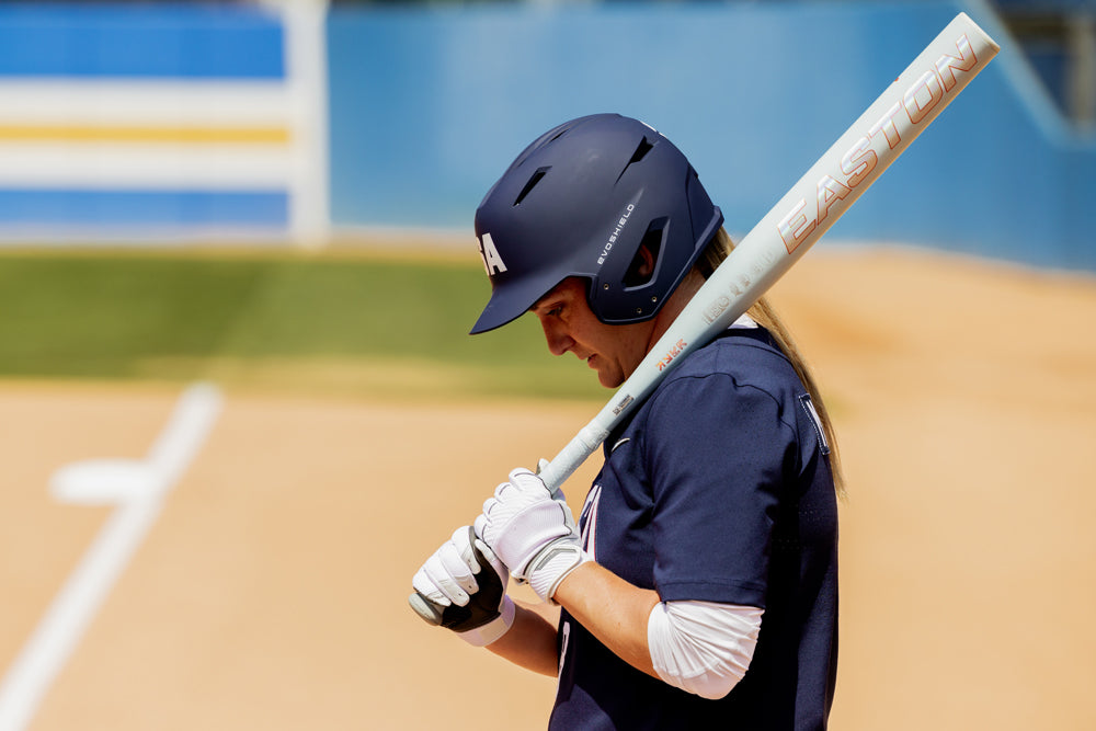 A softball player in a navy uniform and helmet stands at home plate, holding an Easton 2025 Ghost Unlimited (-11) Fastpitch Softball Bat: EFP5GHUL11 (DEMO) over her shoulder on a sunny field.