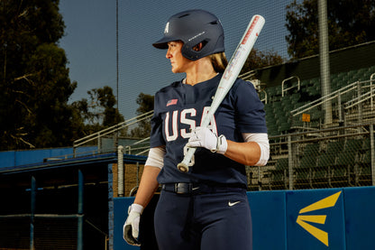 A softball player in a navy blue USA uniform and helmet stands holding the 2025 Easton Ghost Unlimited (-9) Fastpitch Softball Bat (EFP5GHUL9), looking to the side on a field near empty bleachers, fencing, trees, and a blue sky.
