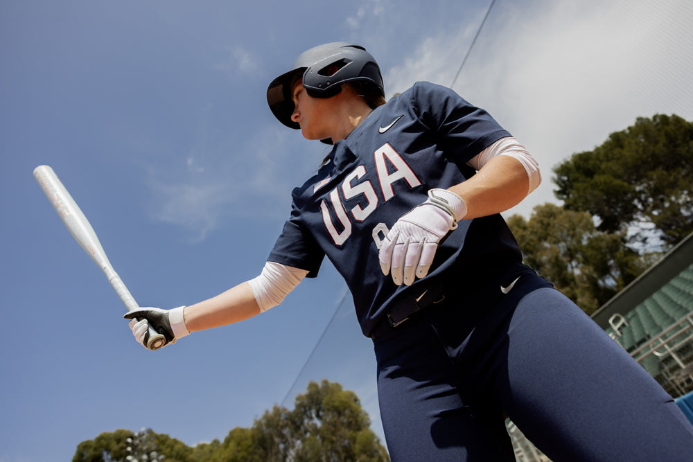 A softball player in a USA uniform and helmet stands ready to bat with the 2025 Easton Ghost Unlimited (-9) Fastpitch Softball Bat (EFP5GHUL9), framed by trees and a blue sky.