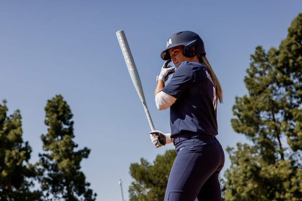 A softball player in a navy uniform grips the 2025 Easton Ghost Unlimited (-9) Fastpitch Softball Bat by Easton, standing ready to bat outdoors amid tall trees and a clear blue sky.