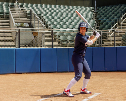 A softball player in a navy USA uniform stands at home plate, gripping the 2025 Easton Ghost Unlimited (-9) Fastpitch Softball Bat (EFP5GHUL9), poised to swing on an empty field with green stadium seats in the background.