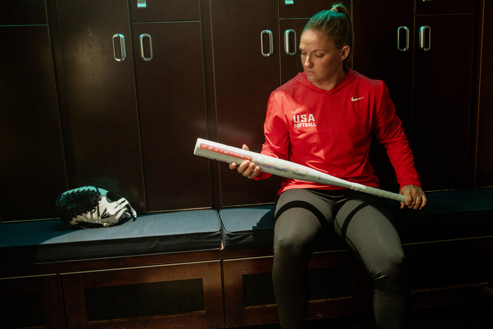 A softball player in a red USA Softball shirt sits on a locker room bench, looking at her 2025 Easton Ghost Unlimited (-10) Fastpitch Softball Bat (EFP5GHUL10) by Easton, with a softball glove resting beside her.