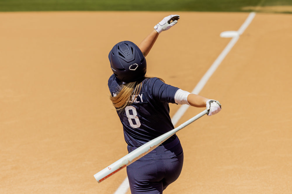 A softball player in a navy uniform and helmet (#8) swings the 2025 Easton Ghost Unlimited (-9) Fastpitch Softball Bat (EFP5GHUL9) by Easton on a sandy field near the foul line, with one arm raised and head turned upward.