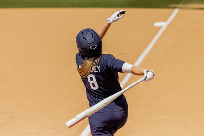 A softball player in a navy uniform and helmet (#8) swings the 2025 Easton Ghost Unlimited (-9) Fastpitch Softball Bat (EFP5GHUL9) by Easton on a sandy field near the foul line, with one arm raised and head turned upward.
