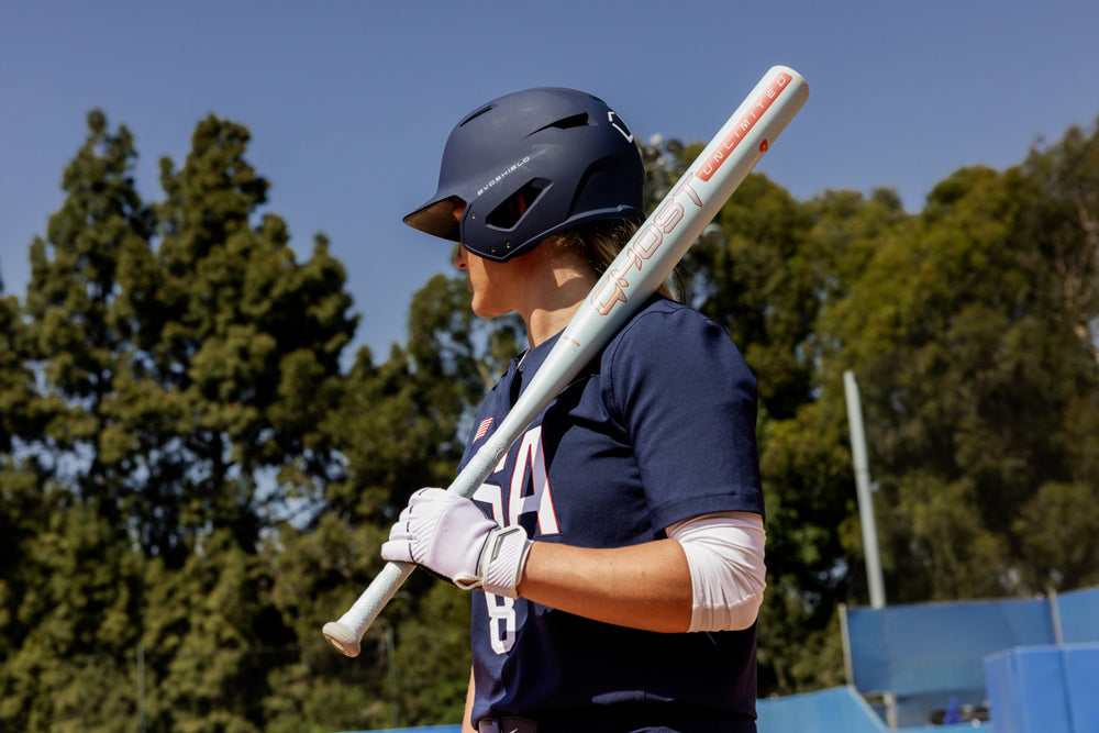 A softball player in a navy #8 uniform holds the 2025 Easton Ghost Unlimited (-9) Fastpitch Softball Bat (EFP5GHUL9) over her shoulder while standing outdoors near trees and a blue sky.