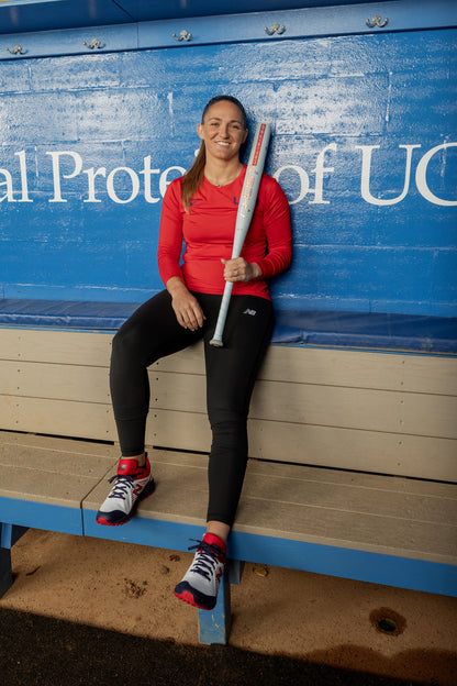 A woman in a red long-sleeve shirt and black pants sits on a bench in a blue dugout, smiling at the camera while holding the 2025 Easton Ghost Unlimited (-10) Fastpitch Softball Bat (EFP5GHUL10) by Easton.