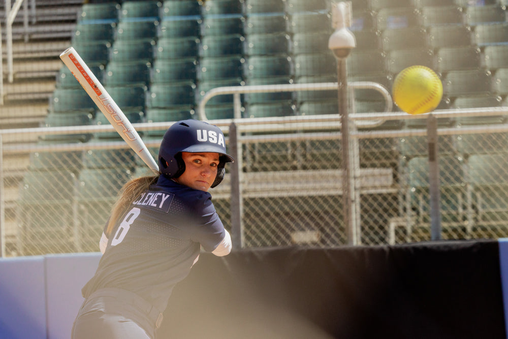 A softball player in a navy USA uniform and helmet readies to swing her 2025 Easton Ghost Unlimited (-11) Fastpitch Softball Bat (EFP5GHUL11 DEMO), eyes locked on the yellow ball, with empty stadium seats behind her.