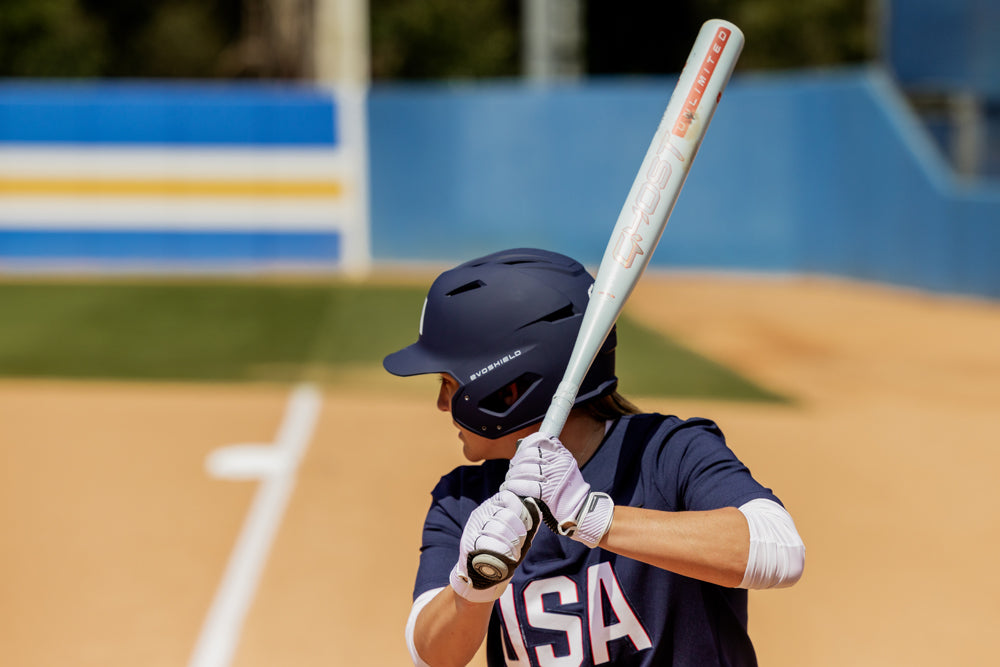 A softball player in a navy helmet and “USA” jersey grips the 2025 Easton Ghost Unlimited (-9) Fastpitch Softball Bat (EFP5GHUL9) by Easton, ready to bat as she faces the pitcher on a field with visible dirt and an outfield fence.
