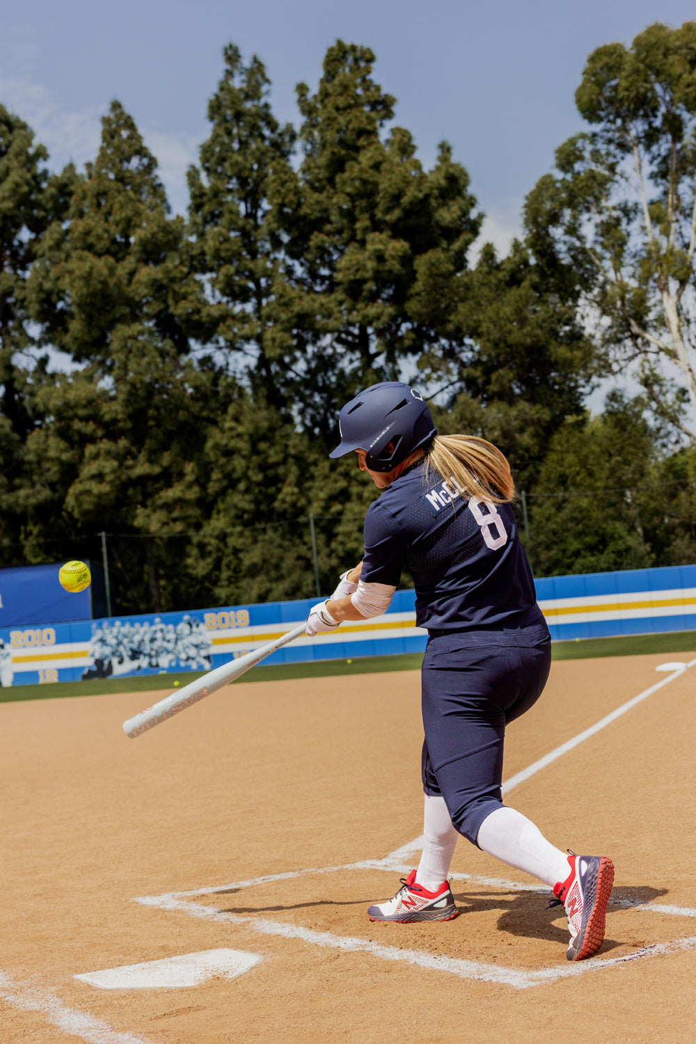 A softball player in navy uniform and helmet swings the 2025 Easton Ghost Unlimited (-9) Fastpitch Softball Bat (EFP5GHUL9), making contact with a yellow softball on a sunny day. Trees and a blue-and-yellow outfield fence are visible behind her.