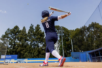 A softball player in a navy uniform with the number 8 swings the 2025 Easton Ghost Unlimited (-9) Fastpitch Softball Bat (EFP5GHUL9) on an outdoor field, with trees and blue stadium seats visible in the background.