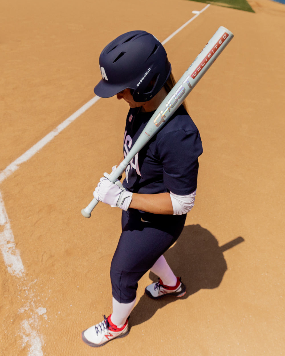 A softball player in a navy uniform and helmet stands on a sandy field near the baseline, holding an Easton 2025 Ghost Unlimited (-9) Fastpitch Softball Bat (EFP5GHUL9) over their shoulder and looking down.