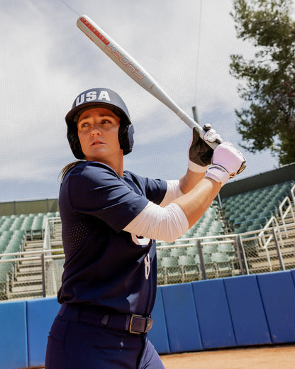 A softball player in a navy USA uniform and helmet grips the 2025 Easton Ghost Unlimited (-11) Fastpitch Softball Bat: EFP5GHUL11 (DEMO), ready to swing at home plate, with empty green bleachers and a blue fence behind.
