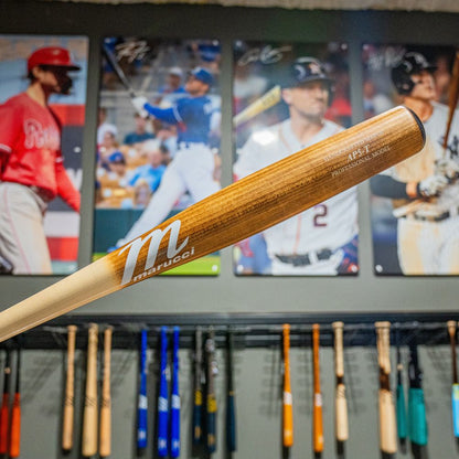A Marucci AP5 Torpedo Pro Model Maple Wood Bat (MVE5AP5T) is held in front of a wall with baseball player posters and a rack of bats, ideal for power hitters seeking premium performance.