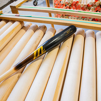 A Marucci CB15 Torpedo Pro Model Maple Wood Bat (MVE5CB15T) with yellow lettering rests on unfinished wooden bats, with more stacked bats in the background.