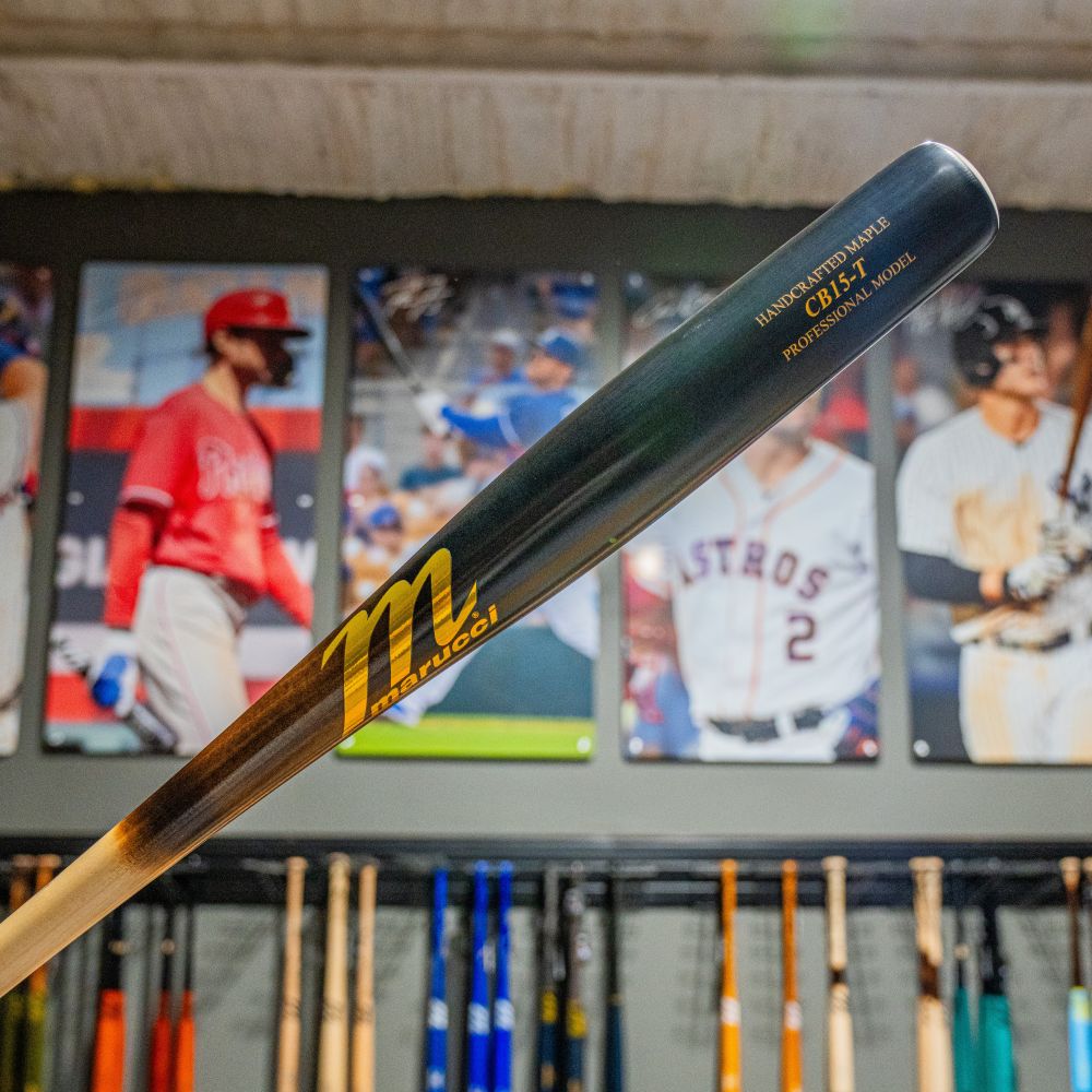 A close-up of a Marucci CB15 Torpedo Pro Model Maple Wood Bat: MVE5CB15T with gold lettering, held in front of a wall with baseball player photos and a rack of end-loaded bats below.