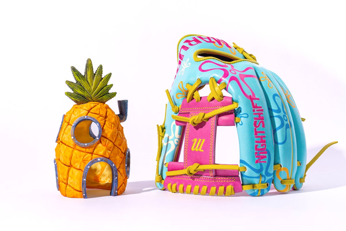 The Marucci NightShift Robert Sponge 11.75" Baseball Glove (MFGNTSHFT-0407) in blue, pink, and yellow sits next to a pineapple-shaped house with windows and green leaves, both displayed on a white background.