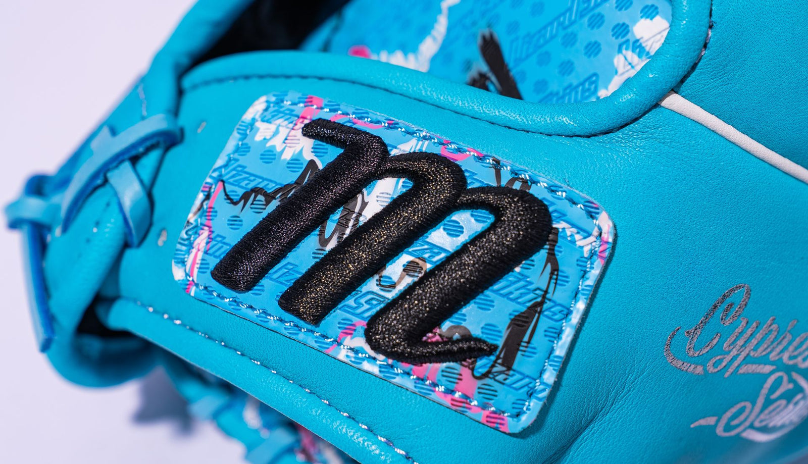 Close-up of a bright blue baseball glove featuring a bold black embroidered M on a patterned blue patch, with some white and pink accents visible in the gloves design.