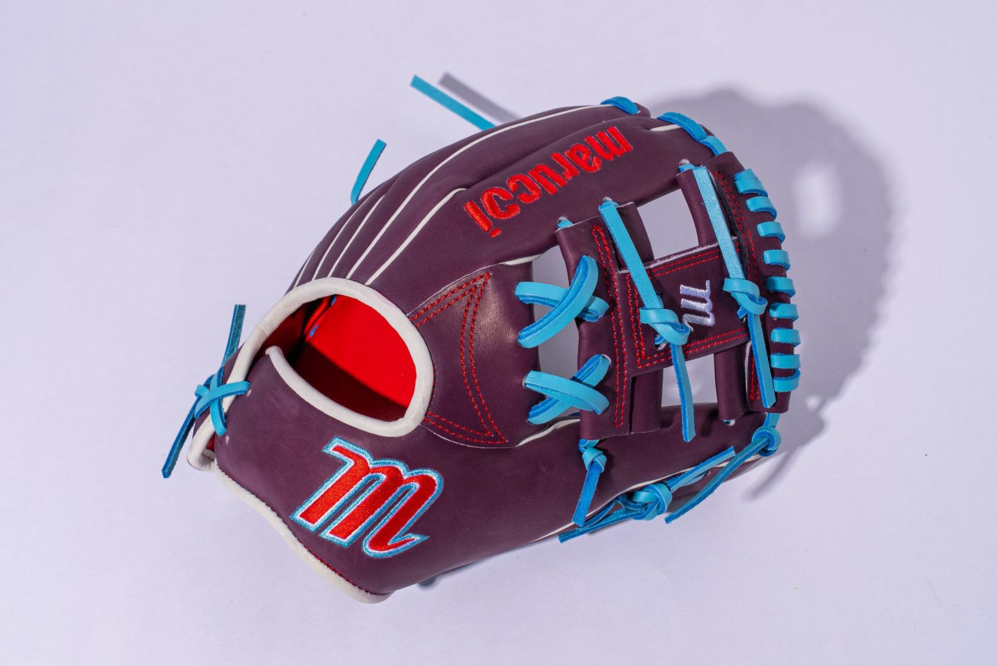 The Marucci NightShift Jetski 11.5" Baseball Glove (MFGNTSHFT-0403), featuring maroon and red Japanese-tanned steerhide, blue laces, and a stylized M logo, is displayed on a light purple background.