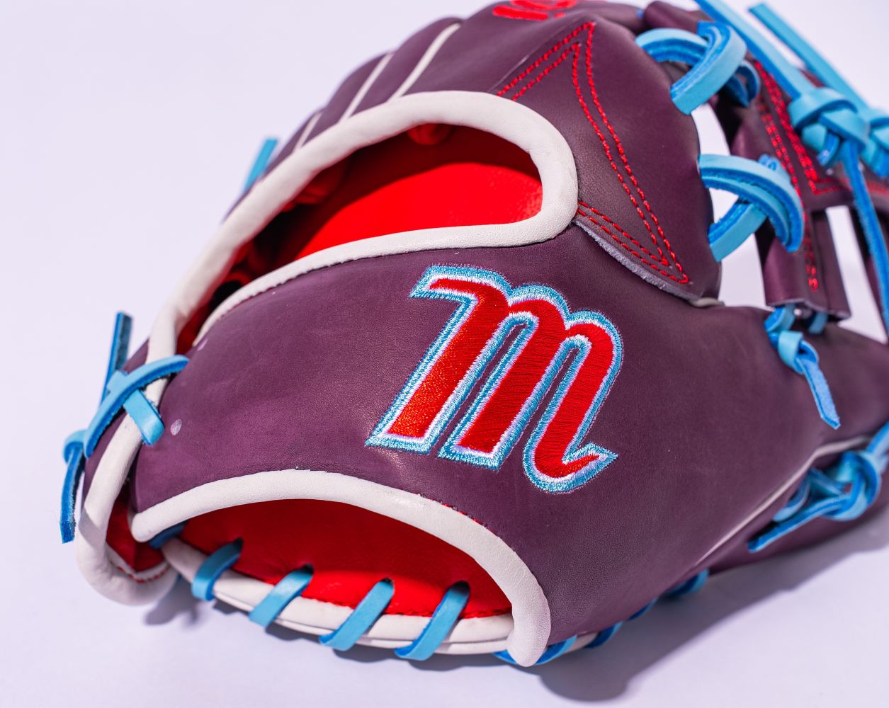 A close-up of the Marucci NightShift Jetski 11.5" Baseball Glove (MFGNTSHFT-0403), crafted from Japanese-tanned steerhide in purple with blue laces, red stitching, a red interior, and a red and white embroidered "M" on the outer side.