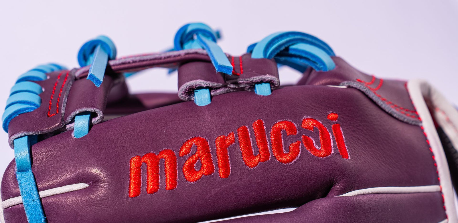 Close-up of a purple Marucci baseball glove with blue laces and red stitching, featuring the brand name marucci embroidered in red on the leather.