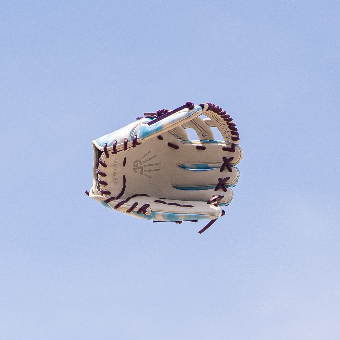 The Marucci NightShift Cloudy 11.75" Baseball Glove (MFGNTSHFT-0402) is shown mid-air, featuring purple laces and light blue accents, crafted from Japanese-tanned steerhide by Marucci, set against a clear blue sky.