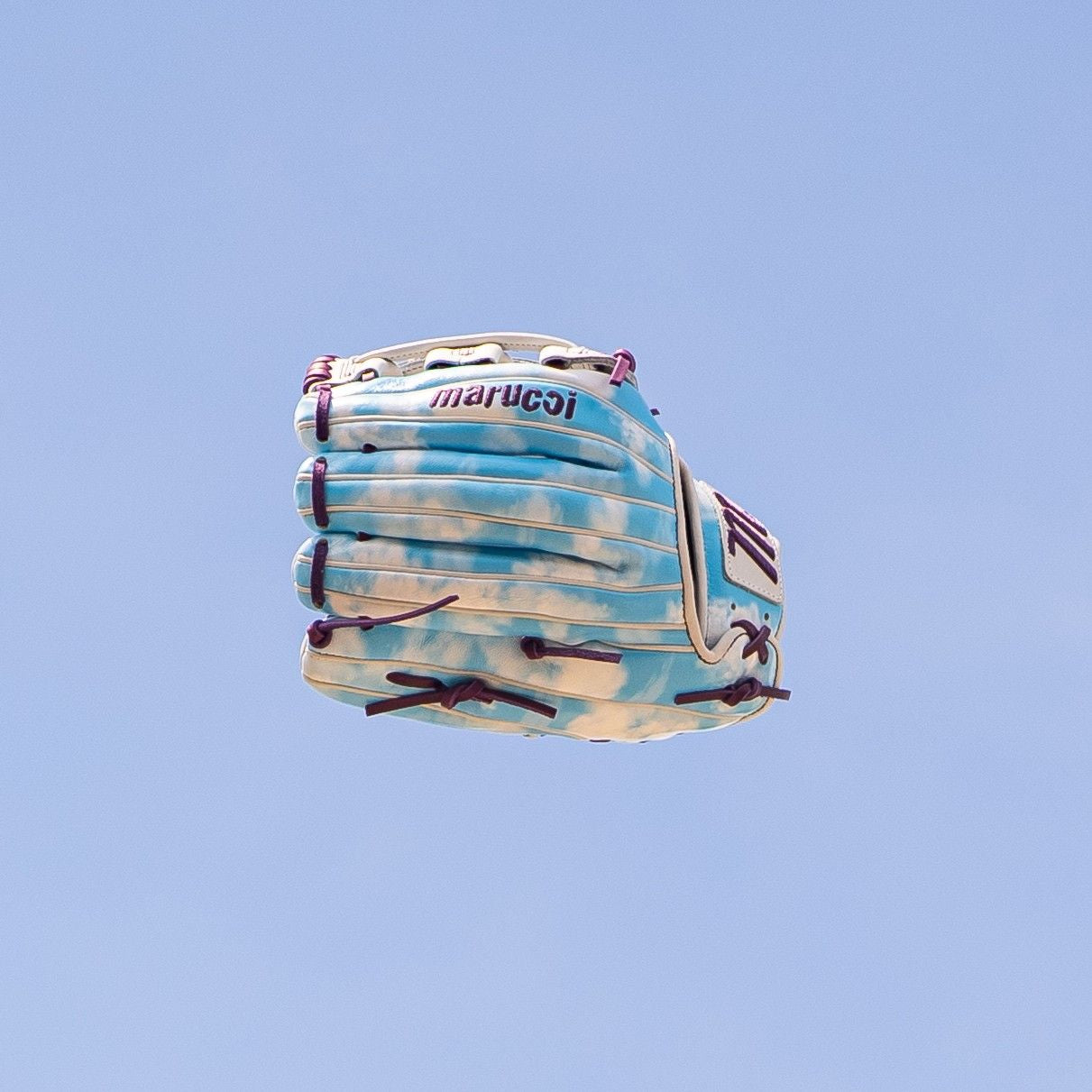 The Marucci NightShift Cloudy 11.75" Baseball Glove (MFGNTSHFT-0402) floats in the air, its blue and white Japanese-tanned steerhide and purple lacing showcased against a clear sky.