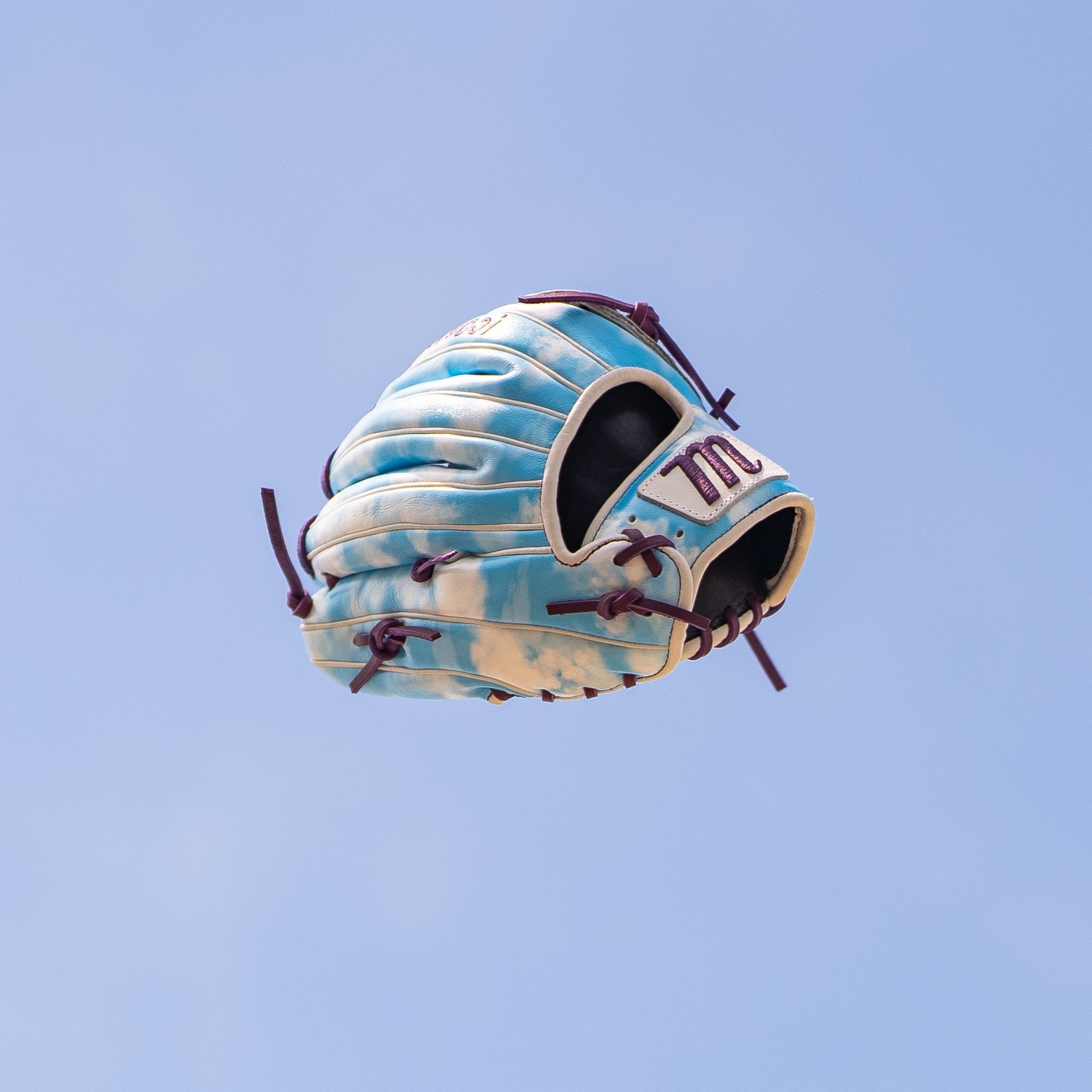 The Marucci NightShift Cloudy 11.75" Baseball Glove (MFGNTSHFT-0402) features Japanese-tanned steerhide in light blue and white leather with maroon laces, standing out vividly against a clear blue sky.