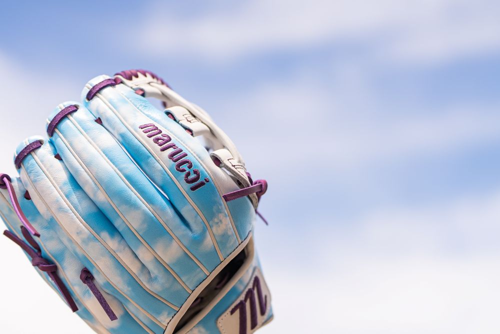 A close-up of a Marucci NightShift Cloudy 11.75" Baseball Glove (MFGNTSHFT-0402), featuring blue, white, and purple accents and crafted from Japanese-tanned steerhide, held against a bright, partly cloudy sky.