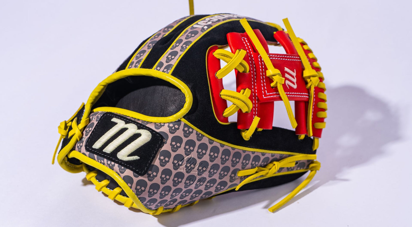 A black baseball glove with yellow laces, red and white accents, and a pattern of gray skulls. The glove features a prominent white M logo near the thumb area.