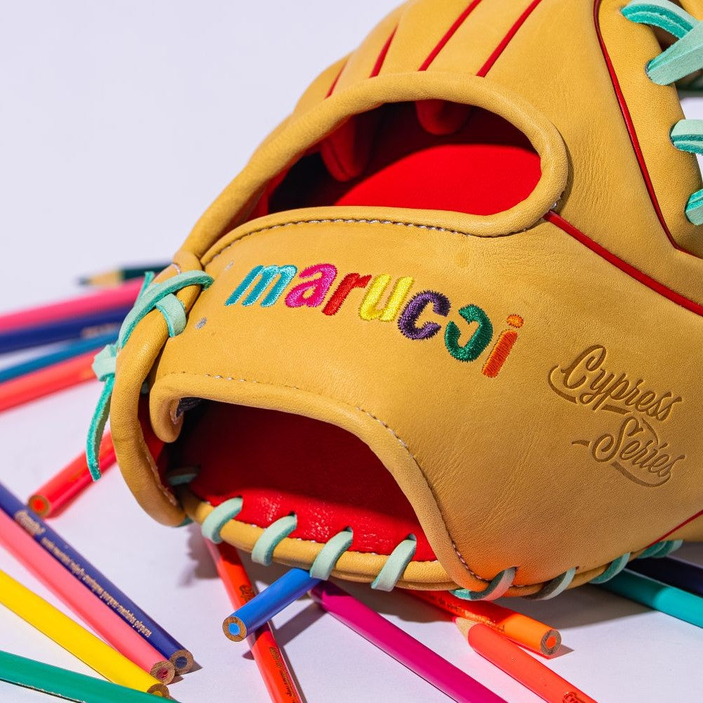 A tan Marucci Cypress Series baseball glove with colorful stitching rests on a scattered pile of colored pencils against a white background.
