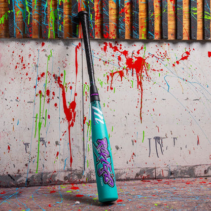A teal Marucci 2026 CAT X RCKLESS Composite (-10) USA Baseball bat with vivid RAVEN graphics leans on a paint-splattered wall with red, green, and blue drips, below a wooden slatted structure.
