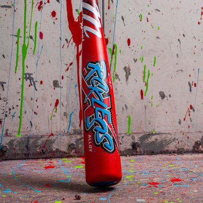 A Marucci 2026 CAT X RCKLESS (-8) USA baseball bat with blue and white graffiti-style text stands upright against a paint-splattered wall, with more colorful splatters scattered across the floor.