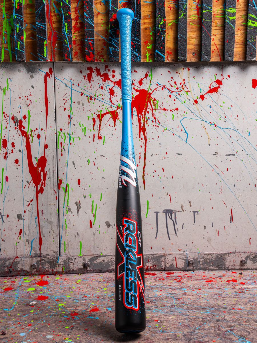 A Marucci 2026 CAT X RCKLESS (-3) BBCOR Baseball Bat with a black handle and blue grip stands upright against a wall with colorful paint splatters and wooden slats. The bat features "Reckless" in bright letters.