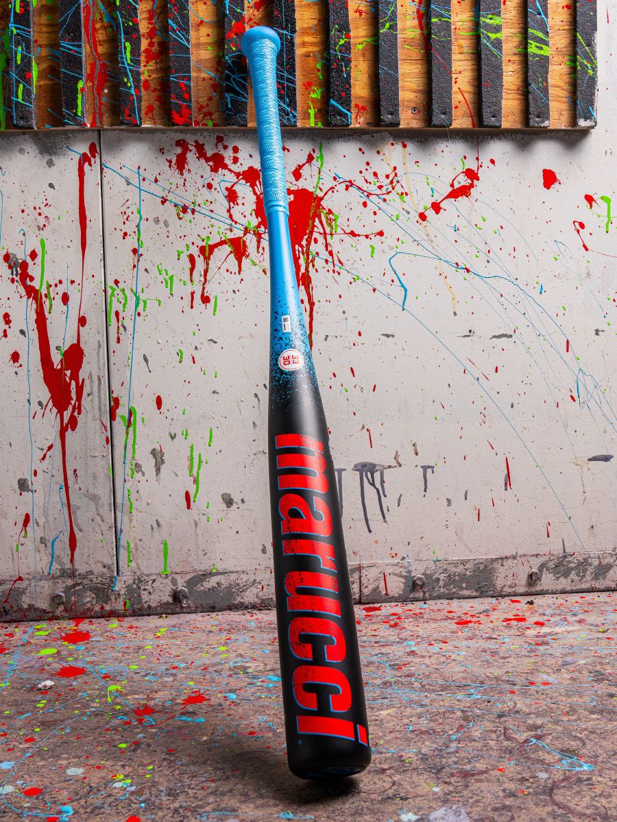 A 2026 Marucci CAT X RCKLESS (-3) BBCOR Baseball Bat (MCBCR) with a blue handle and black barrel featuring red Marucci branding stands upright against a paint-splattered wall, with vertical wooden planks visible at the top.