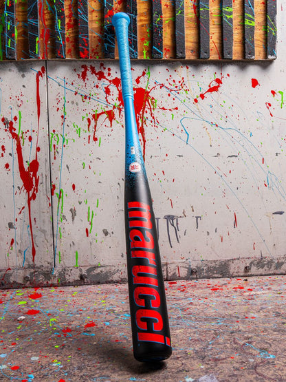 A 2026 Marucci CAT X RCKLESS (-3) BBCOR Baseball Bat (MCBCR) with a blue handle and black barrel featuring red Marucci branding stands upright against a paint-splattered wall, with vertical wooden planks visible at the top.