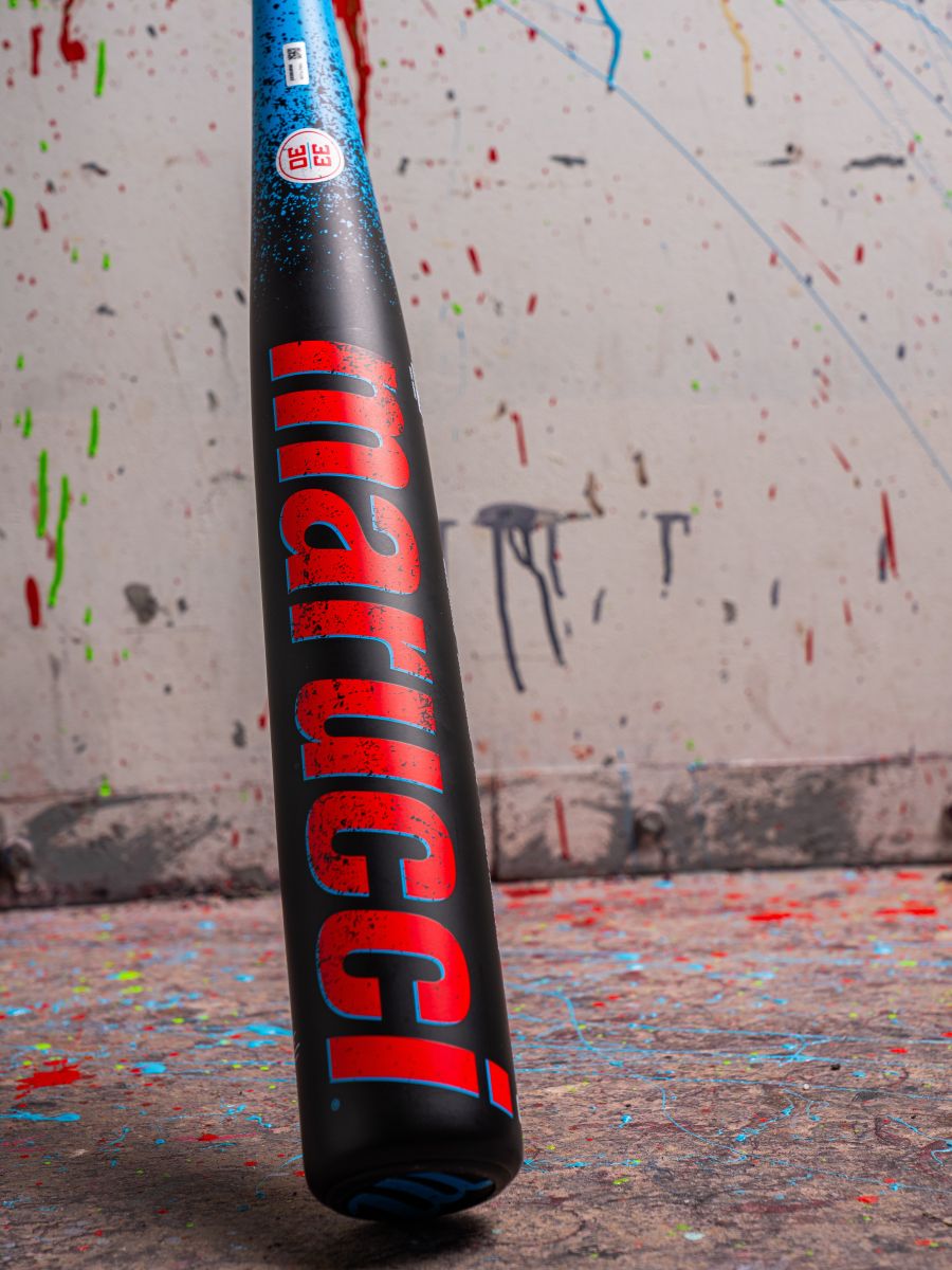 A 2026 Marucci CAT X RCKLESS (-3) BBCOR Baseball Bat (MCBCR) stands upright with bold red and blue branding on a paint-splattered floor, leaning against a paint-covered wall.