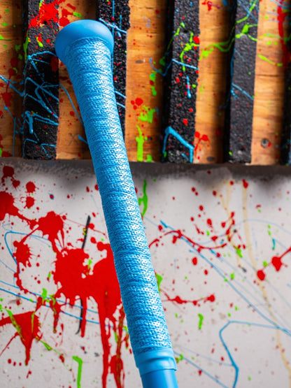 A close-up of the Marucci CAT X RCKLESS (-3) BBCOR Baseball Bat (MCBCR) handle in blue alloy is set against a wall with splattered red, blue, and green paint and wooden slats.