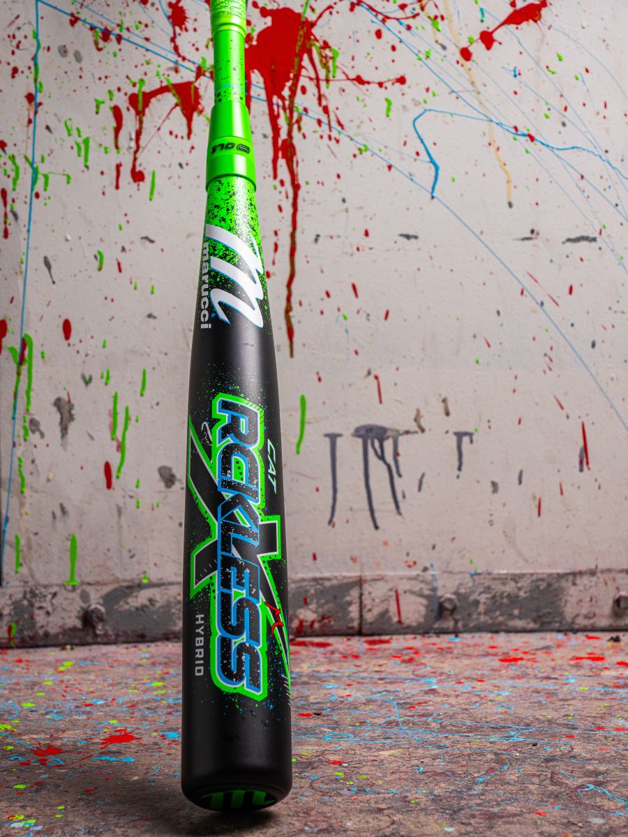 A neon green and black Marucci 2026 CAT X RCKLESS Hybrid (-3) BBCOR Baseball Bat (MCBCRH DEMO) stands upright on a paint-splattered floor, with a wall behind it covered in vibrant drips and splatters.