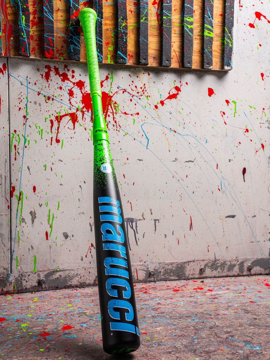 A 2026 Marucci CAT X RCKLESS Hybrid (-3) BBCOR Baseball Bat (Demo), with a bright green handle, black barrel, and blue Marucci logo, stands upright against walls and floors splattered with colorful paint.