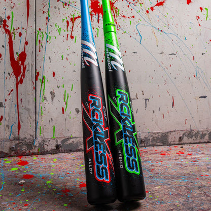 Two Marucci 2026 CAT X RCKLESS Hybrid (-3) BBCOR baseball bats, one blue and one green, stand upright against a wall splattered with red, blue, and green paint; vibrant splatters cover the floor.