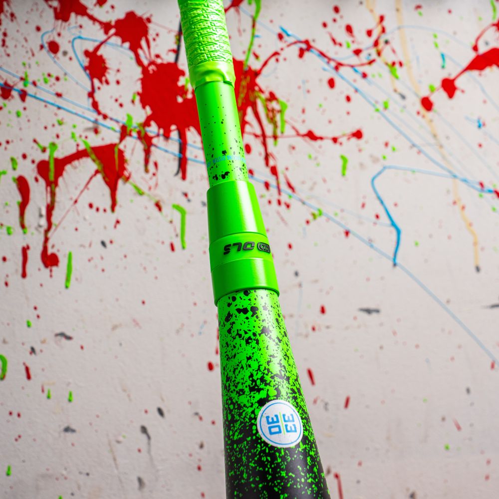 A close-up of the Marucci 2026 CAT X RCKLESS Hybrid (-3) BBCOR Baseball Bat (DEMO), featuring green and black paint splatters, is held upright before a wall covered with red, blue, and green paint streaks.