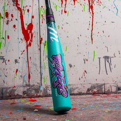 The 2026 Marucci CAT X RCKLESS Composite (-10) USA Baseball Bat stands upright on a paint-splattered floor, its bold Ruckus graphics and turquoise design contrasting against a white wall splattered with red, green, and blue paint.