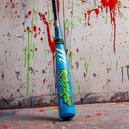 A 2026 Marucci CAT X RCKLESS Hybrid (-11) USA Baseball Bat stands upright on a splattered floor, with red, green, and blue paint covering the wall and ground behind it.