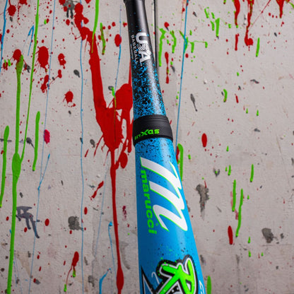 A close-up of a blue Marucci CAT X RCKLESS Hybrid (-11) 2 5/8" USA Baseball Bat (MSBCRH11USA) with neon green lettering, held upright against a white wall splattered with red, green, and blue paint.