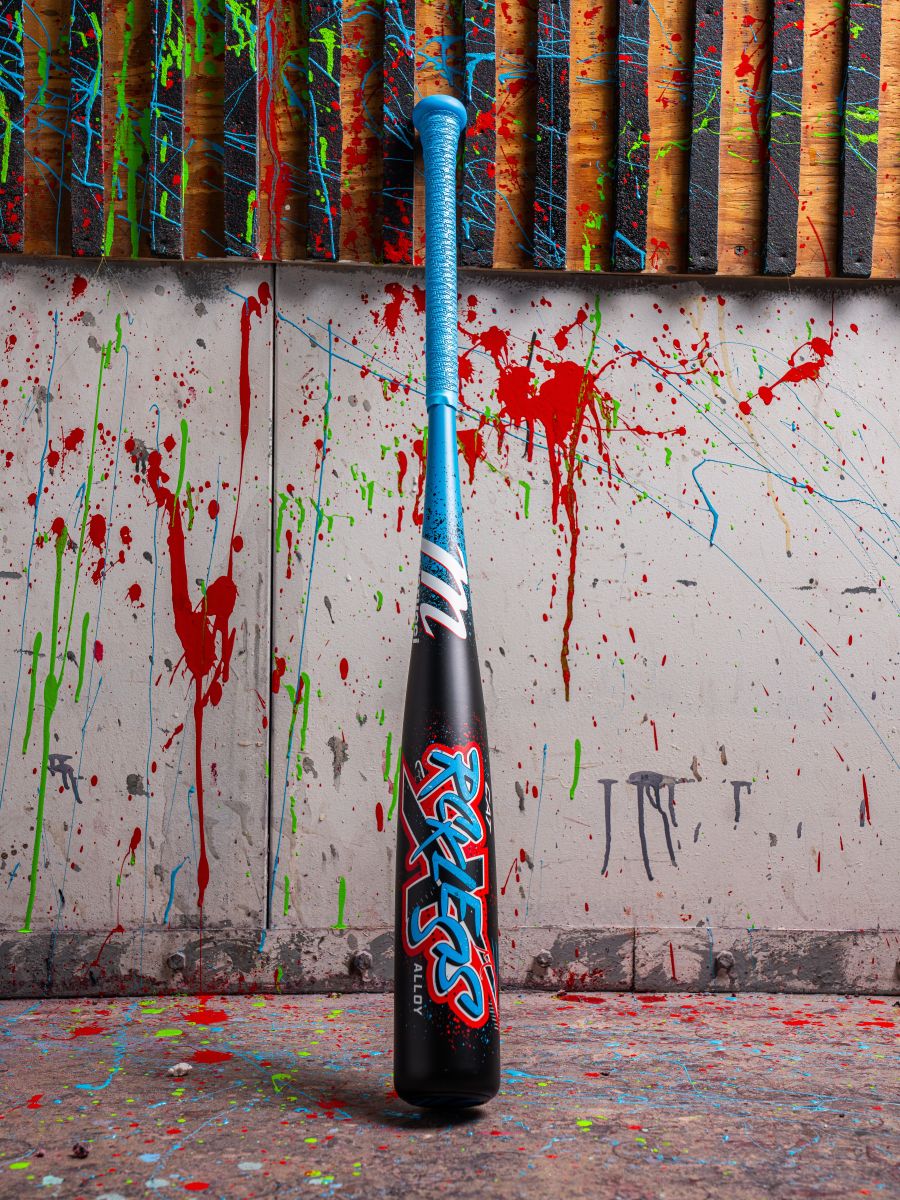 The 2026 Marucci CAT X RCKLESS (-8) USSSA Baseball Bat, featuring a blue handle and graffiti-style text, stands upright against a wall covered in splashes of red, green, and blue paint, with vibrant colors scattered across the floor.