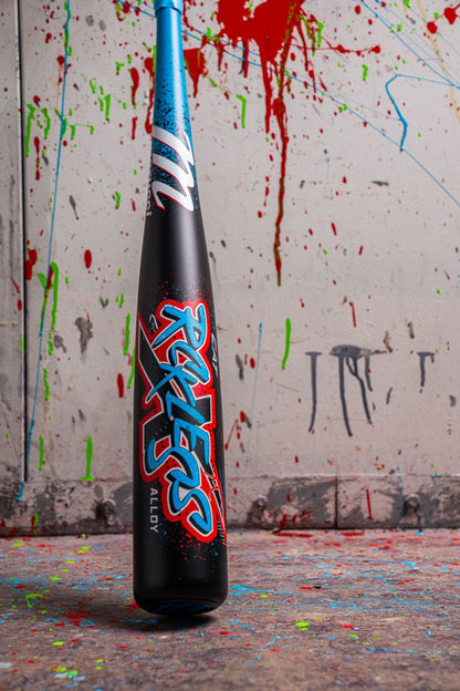 A black and blue Marucci CAT X RCKLESS (-8) 2 3/4" USSSA Baseball Bat (MSBCR8) stands upright on a paint-splattered floor, set against a wall streaked with bold red, blue, and green paint.