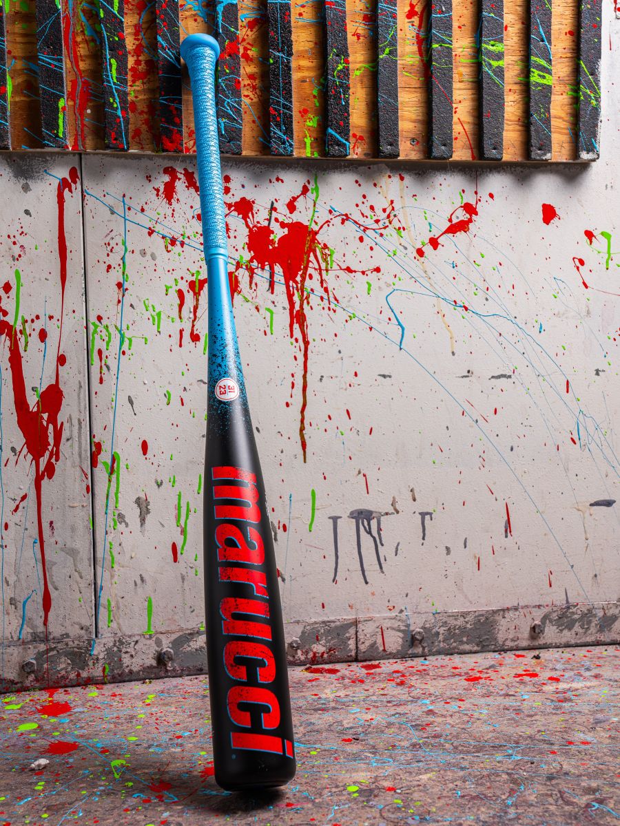 A 2026 Marucci CAT X RCKLESS (-8) 2 3/4" USSSA Baseball Bat (MSBCR8) with a blue handle and black barrel featuring the red Marucci logo stands upright against a paint-splattered wall with wooden panels in the background.