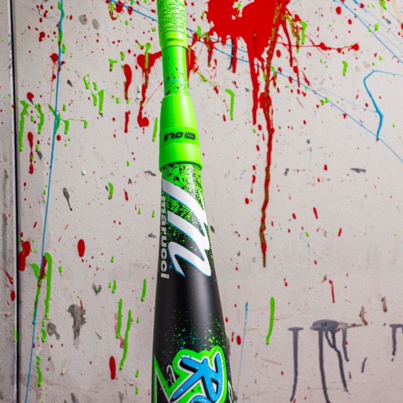 A Marucci CAT X RCKLESS Hybrid (-5) 2 3/4" USSSA Baseball Bat (MSBCRH5), in neon green and black, stands upright before a wall splattered with red, blue, and gray paint. Bold Marucci branding and striking graphics are featured.
