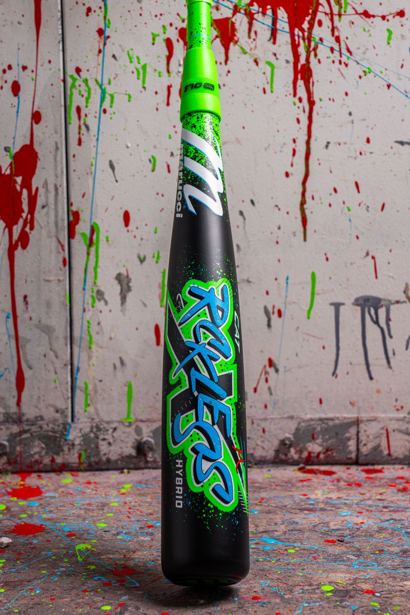 A 2026 Marucci CAT X RCKLESS Hybrid (-5) USSSA baseball bat stands upright on a splattered paint floor, vibrant against a wall of colorful paint drips, highlighting its bold design and Marucci branding.